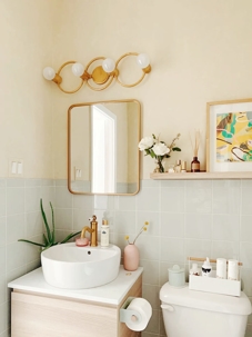 Stylish bathroom with light wood vanity, round vessel sink, brass fixtures, soft green tile backsplash, and minimalist decor including plants, art, and modern lighting.